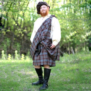 Puede incluir: Un atuendo tradicional escocés de kilt. El conjunto incluye una boina marrón, una camisa color crema, un kilt de tartán en tonos grises, azules y burdeos, una faja a juego, un sporran de cuero negro y botas altas negras. El atuendo se usa al aire libre.