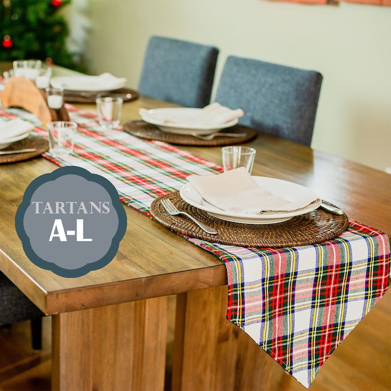 May include: A red, white, and blue plaid table runner with a woven placemat and white dinner plate on a wooden table. The table runner is labeled "TARTANS A-L".