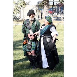May include: A man and a woman wearing traditional Scottish kilts and clothing. The man is wearing a green kilt with a black sporran and a green jacket. The woman is wearing a black kilt with a white apron and a green jacket. They are both standing on a grassy field.