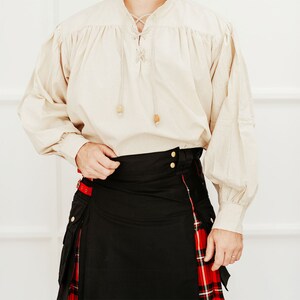 May include: A white linen shirt with a lace-up neckline and long sleeves, worn with a black kilt featuring a red and black tartan trim.