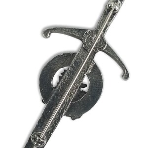 May include: Silver-toned kilt pin featuring a sword design with a circular emblem. The pin has a textured finish and a safety clasp.