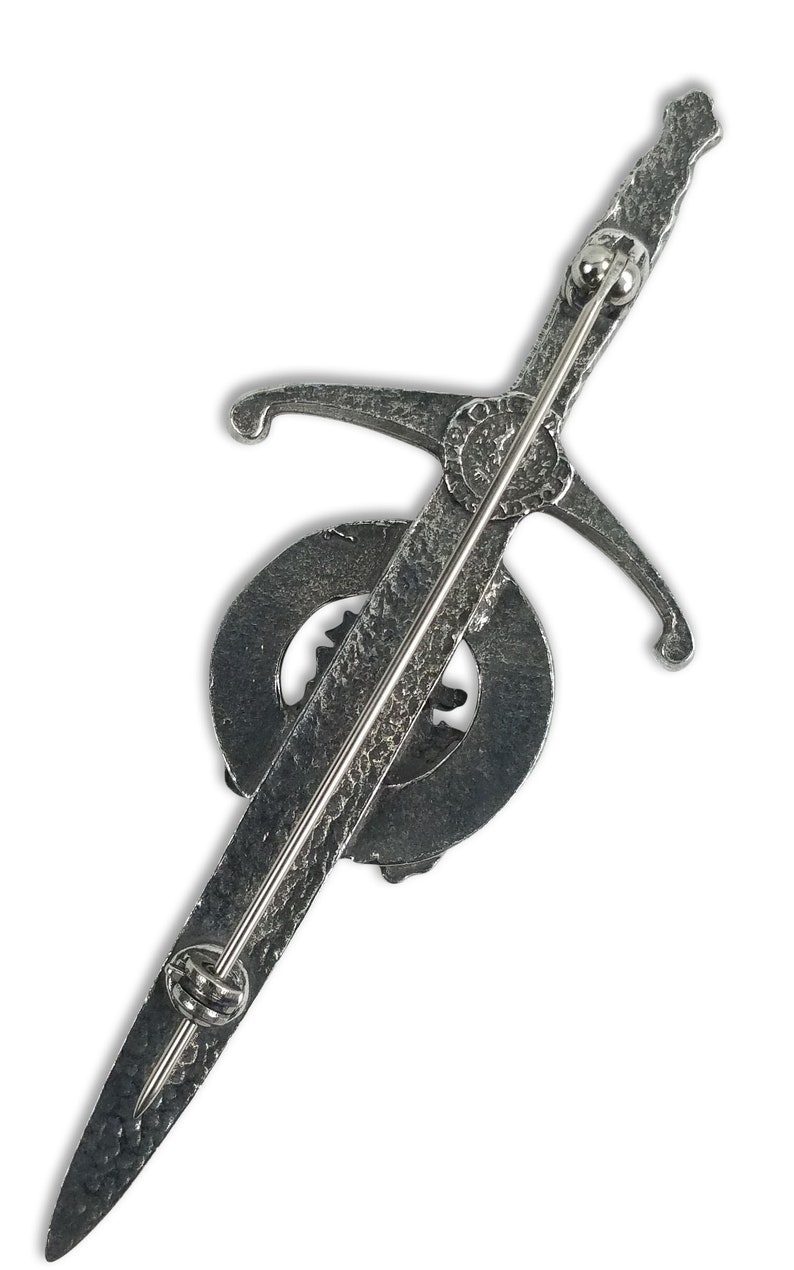 May include: Silver-toned kilt pin featuring a sword design with a circular emblem. The pin has a textured finish and a safety clasp.
