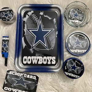 May include: A Dallas Cowboys themed smoking set with a silver tray, a glass ashtray, three small containers, and a lighter. The tray has a blue and white design with the Cowboys logo and the word "COWBOYS" printed on it. The ashtray and containers also feature the Cowboys logo.