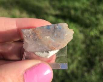 May include: A small, rough, white and blue crystal with a few brown flecks. The crystal is sitting on a clear glass square.