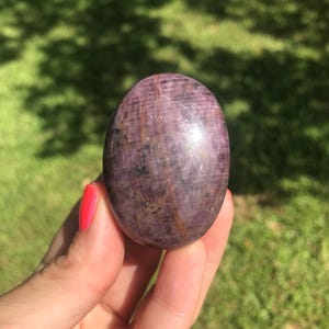 May include: A smooth, polished oval-shaped stone with a deep purple and magenta hue. The surface features natural dark inclusions, subtle linear striations, and reflective highlights that emphasize its rounded, symmetrical form and texture.