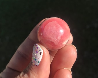 May include: A polished, translucent pink sphere, held in a hand. The sphere has a marbled appearance with lighter pink and white areas. The background is a blurred green and dark area. The fingernail has a white base with colorful glitter.