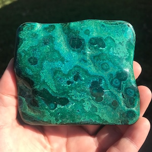 May include: A polished, rectangular stone with a vibrant green and teal color palette. The stone's surface features a unique pattern of swirls and spots, creating a natural, organic aesthetic. The stone is held in a hand, showcasing its size and texture.