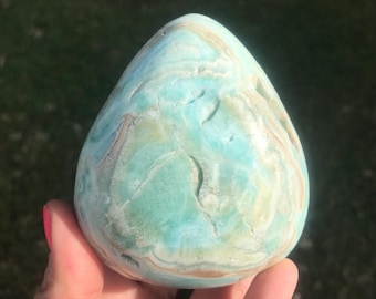May include: A teardrop-shaped stone with a blend of blue, tan, and white hues. The polished surface reveals a swirling pattern, creating a marbled effect. The stone is held in a hand, showcasing its smooth texture and natural beauty.
