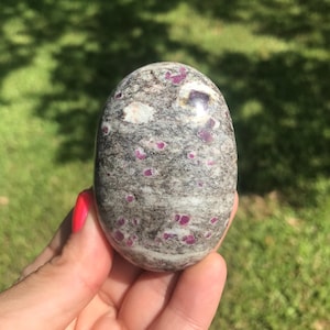 May include: A smooth, oval-shaped polished stone featuring a mottled gray and white base with scattered, vibrant magenta-pink inclusions throughout the surface. The stone has a glossy finish and displays a natural, varied mineral texture.