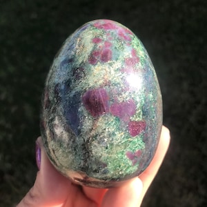 May include: A polished, egg-shaped gemstone with a mix of colors. The stone features a base of green and blue-grey hues, with vibrant red and purple inclusions scattered throughout. The surface is smooth and reflective, catching the light.