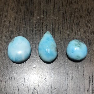 May include: Three polished larimar gemstones in different shapes: an oval, a teardrop, and a round cabochon. The stones display a light blue color with subtle patterns, set against a dark wood grain surface.