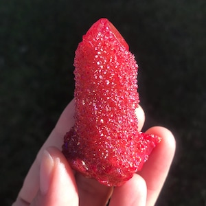 May include: A vibrant red crystal cluster with a sparkling, textured surface. The crystal has a pointed top and a rough, irregular shape. The color is a deep, saturated red with hints of iridescent shimmer, suggesting a druzy or coated finish.