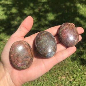 May include: Three smooth, polished oval stones with a mottled appearance. The stones feature a blend of earthy brown, deep red, and muted blue-green tones with a glossy, reflective surface that catches the light, showcasing natural mineral textures.