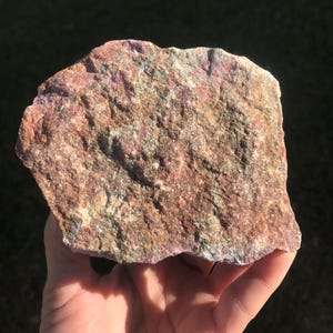May include: A rough-hewn, reddish-brown rock with patches of green and gray. The natural stone has a textured surface and is held in a hand, with a dark background.