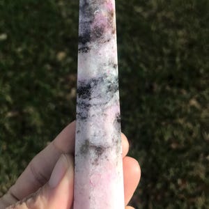 May include: A polished crystal obelisk with a pointed top. The stone features a blend of pink, black, and gray hues. The crystal is held in a hand against a blurred green background.