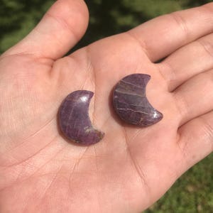 May include: Two crescent moon-shaped carvings made of a deep purple and magenta mineral with visible natural striations and a polished, smooth surface. These small, curved stone pieces feature distinct color variations and a glossy finish.