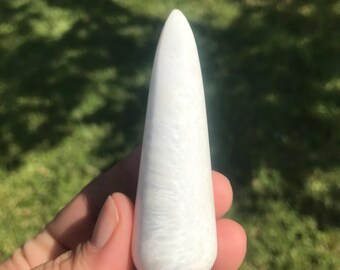 May include: A white, elongated crystal wand with a rounded base and a tapered top. The wand has a marbled appearance with subtle gray veining throughout. The object is held in a hand against a blurred green background.