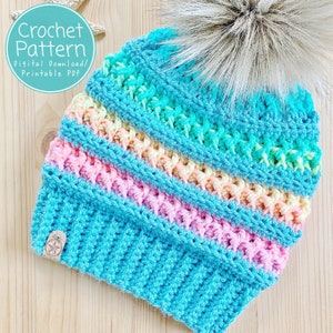 May include: A crocheted hat with a rainbow stripe pattern in shades of pink, yellow, and blue. The hat has a fluffy white pompom on top and a brown leather tag with a star.