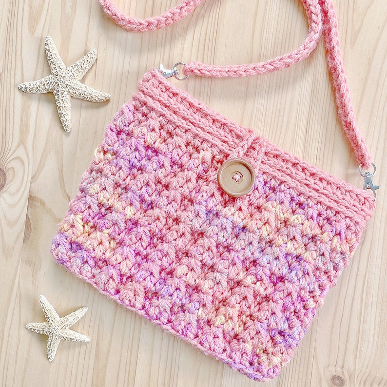 K&ouml;nnte beinhalten: Eine rosa geh&auml;kelte Umh&auml;ngetasche mit braunem Knopfverschluss. Die Tasche hat einen hellrosa, gelben und violetten Ombre-Effekt.