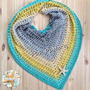 Puede incluir: Un chal de crochet de color turquesa, amarillo y gris con un adorno de estrella de mar de crochet. El chal está colocado sobre una superficie de madera. El texto "PDF Pattern" es visible en el chal.