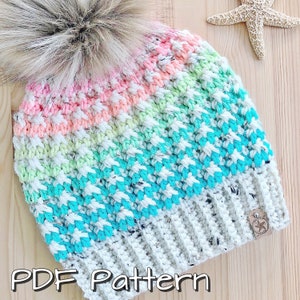 May include: A colourful crochet beanie hat with a white pom pom. The hat is made with a textured stitch and features a rainbow ombre colour pattern. The hat has a brown leather tag with a star on it. The text "PDF Pattern" is written below the hat.