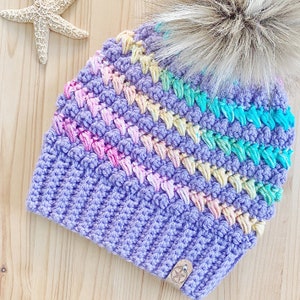K&ouml;nnte beinhalten: Eine lilafarbene H&auml;kelm&uuml;tze mit einem Regenbogenstreifenmuster und einem flauschigen wei&szlig;en Bommel oben.