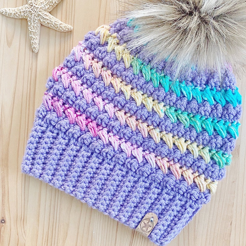 K&ouml;nnte beinhalten: Eine lilafarbene H&auml;kelm&uuml;tze mit einem Regenbogenstreifenmuster und einem flauschigen wei&szlig;en Bommel oben.
