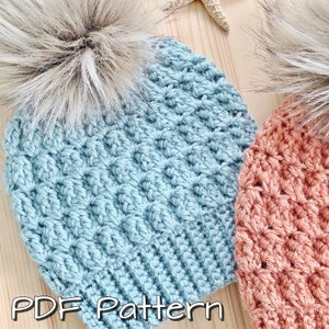 May include: Two crocheted hats with faux fur pom poms. One hat is light blue and the other is a peachy orange colour. The hats are on a wooden surface with the text "PDF Pattern" at the bottom of the image.
