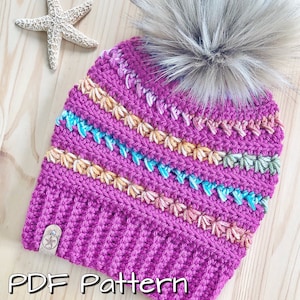 May include: A magenta crochet hat with a faux fur pom-pom. The hat has bands of gold, turquoise, and green. The text "PDF Pattern" is at the bottom. A starfish is in the background. The hat is a handmade item.