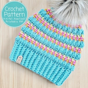 May include: A crochet hat pattern featuring a blue base with colourful stripes of yellow, pink and orange. The hat has a fluffy white pompom on top. The pattern is a digital download and printable PDF.