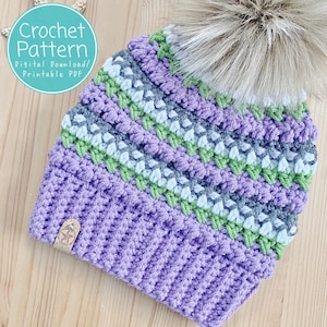 May include: Purple, green, white and grey crochet beanie with a faux fur pom pom. The beanie has a striped pattern and the text "Crochet Pattern Digital Download/ Printable PDF" is on a teal circle.