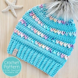 Op de afbeelding: Een turquoise gehaakte muts met een imitatiebont pompon. De hoed heeft een gestructureerd steekpatroon met pastelkleuraccenten. Een klein leren label is aan de zijkant bevestigd. De tekst "Crochet Pattern" staat in een cirkel.