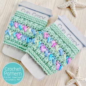 Könnte beinhalten: Zwei grüne gehäkelte Becherwärmer mit einem bunten Garnmuster. Die Becherwärmer befinden sich auf braunen Papierkarten mit dem Text "Cup Cozy" und "Made with love".