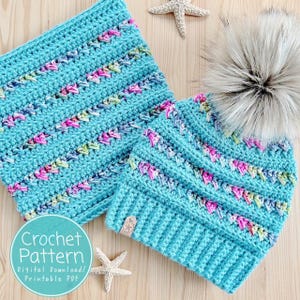 Pode incluir: Um conjunto de gorro e gola de crochê turquesa com um padrão floral em rosa, verde e branco. O gorro apresenta um grande pompom cinza fofo. O texto "Crochet Pattern" é visível em um gráfico circular.