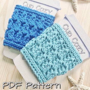 May include: Two crocheted cup cozies, one blue and one teal, with the text "Cup Cozy" printed on the tags. The cozies are made with a textured stitch pattern.  PDF Pattern.