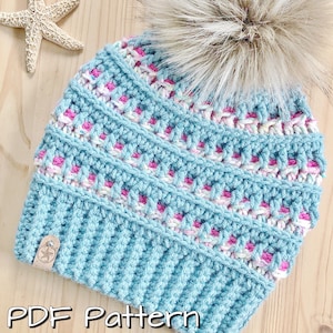 May include: A blue and pink crocheted beanie with a fluffy white pom pom. The beanie has a striped pattern and a small tag with a star on it. PDF Pattern.