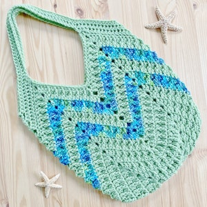 K&ouml;nnte beinhalten: Eine gr&uuml;ne geh&auml;kelte Tragetasche mit einem blauen und gr&uuml;nen geometrischen Muster. Die Tasche hat eine breite, offene Oberseite und einen einzigen Riemen.