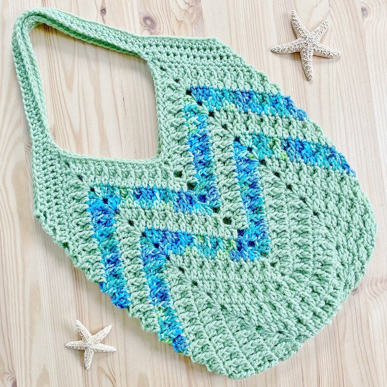K&ouml;nnte beinhalten: Eine gr&uuml;ne geh&auml;kelte Tragetasche mit einem blauen und gr&uuml;nen geometrischen Muster. Die Tasche hat eine breite, offene Oberseite und einen einzigen Riemen.