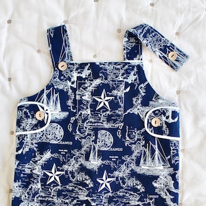 Könnte beinhalten: Marineblaue Baby-Latzhose mit nautischem Kartenmuster mit Segelbooten, Ankern und Sternen. Die Latzhose hat Holzknöpfe, weiße Paspeln und verstellbare Träger. Ein stilvolles und bequemes Outfit für ein Kleinkind.