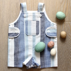 Könnte beinhalten: Ein blau-weiß gestreifter Baby-Strampler mit Knopfdetails und Taschen. Der Strampler ist aus weichem Stoff gefertigt. Dekorative gesprenkelte Eier in Grün, Rosa und Weiß liegen daneben.