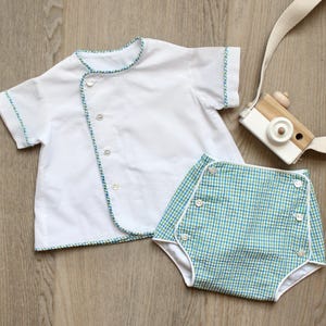 May include: A white short-sleeved shirt with a diagonal button closure and blue and green trim, paired with blue and green gingham bloomers featuring white buttons and white piping. A wooden toy camera with a white strap is also included.