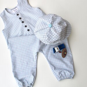 May include: A light blue and white checkered baby outfit set, including a vest with black buttons, matching pants, and a cap. The pants feature a small blue and brown toy truck applique.