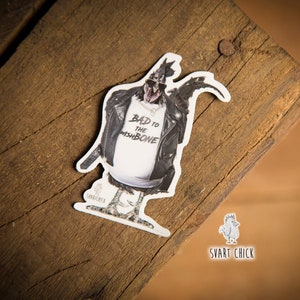 May include: A black and white sticker of a chicken wearing a leather jacket and a white t-shirt that says "Bad to the Wishbone". The sticker is on a wooden surface. The text "Svart Chick" is visible below the sticker.