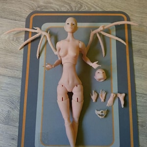 May include: A nude-colored doll with wings, head, hands, feet, and a jawbone. The doll is laying on a gray mat with an orange border.