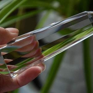 May include: A clear quartz crystal point, also known as a crystal wand, is held in a hand. The crystal is long and slender with a pointed tip. The crystal is reflecting light and has a slight rainbow effect.