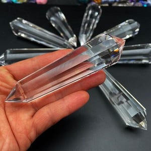 Puede incluir: Una colección de varitas de cristal de cuarzo transparente, algunas con facetas, expuestas sobre una superficie negra. Las varitas son de diferentes longitudes y se sostienen en una mano.