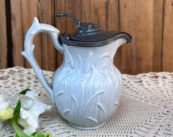 May include: A light blue ceramic pitcher featuring a raised relief pattern of tall grass blades on the body and handle. The pitcher is topped with a dark gray metal hinged lid that includes a small, textured metal finial on the top surface.