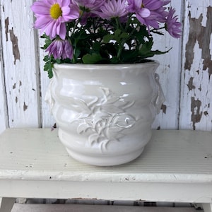 May include: A white ceramic planter with a floral design, featuring a bouquet of light purple flowers.