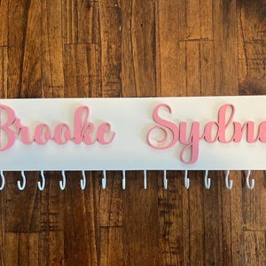 Op de afbeelding: Een witte houten medailles hanger met de namen "Brooke" en "Sydnee" in roze letters. De hanger heeft een rij witte haken.