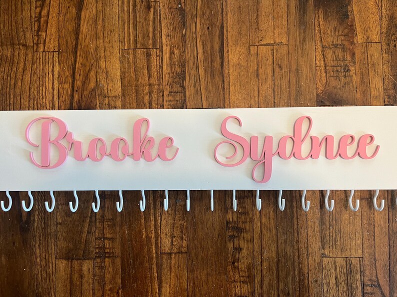 Op de afbeelding: Een witte houten medailles hanger met de namen "Brooke" en "Sydnee" in roze letters. De hanger heeft een rij witte haken.